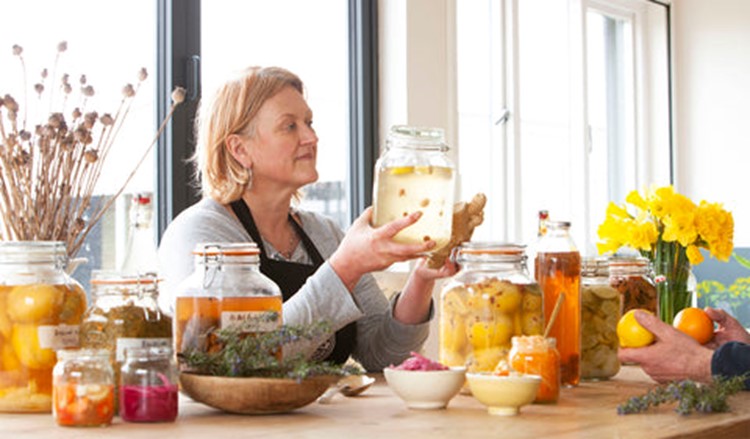 Images shows a woman holding a jar of food with other pickles and ferments in jars around her.